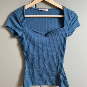 Blå ribbad topp från Anna Field - Supersnygg blå ribbad topp från Anna Field med korta ärmar och en unik v-ringad halsringning. Toppen har figurnära passform och vertikala ränder som ger en cool struktur. Perfekt att styla med jeans eller kjol för en trendig look.
