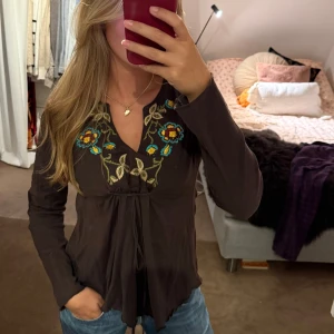 Brun blus med broderade blommor - Supersöt brun blus med broderade blommor i gult, blått och beige vid ringningen. V-ringad med knyt framtill och långa ärmar🌻