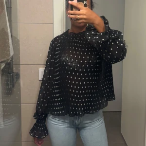 Svart prickig blus med volangärm - Säljer en svart transparent blus med vita prickar och volangdetaljer vid ärmslut och hals. Blusen har lång ärm och en lös, luftig passform som ger en cool och trendig look. Perfekt att styla med jeans eller kjol för en snygg outfit.