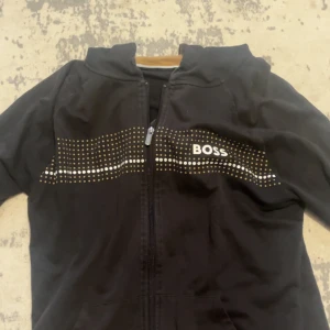 Svart hoodie från BOSS med prickar - Svart hoodie från BOSS i storlek M med dragkedja och huva. Framsidan har ett coolt prickigt mönster i guld och vitt samt BOSS-loggan tryckt över bröstet. Perfekt för dig som gillar streetstyle och vill ha något som sticker ut. Pris kan diskuteras!