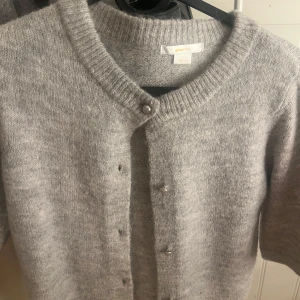 Grå kofta från Gina Tricot - Mysig grå kofta från Gina Tricot med kortare ärmar och pärlknappar framtill. Koftan är stickad och har en rund halsringning med bred ribbad kant. Perfekt för lager-på-lager och enkel att styla till olika looks.