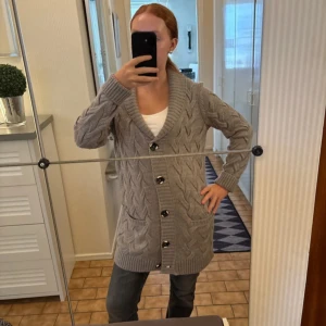 Grå lång kofta med knappar - Säljer en mysig grå kofta med grovstickat flätmönster och stora svarta knappar. Koftan är lång, har två fickor fram och bred krage. Perfekt att slänga på sig när det är kyligt ute. Passar dig som gillar chill och comfy vibes. Nyskick aldrig använd 