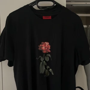 Svart t-shirt med ros från Hugo Boss - Svart t-shirt från Hugo Boss med en stor, detaljerad röd ros tryckt på bröstet. Klassisk rund hals och korta ärmar. Materialet är mjuk bomull som känns skönt mot huden. Perfekt för dig som gillar stilrena plagg med en twist.