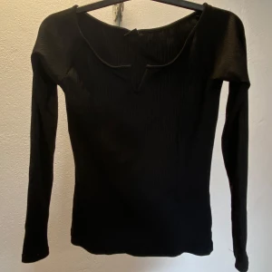 Svart ribbad topp från Gina Tricot - Svart långärmad topp från Gina Tricot i ribbat material. Toppen har en unik v-ringad detalj vid halsen och är figurnära i modellen. Perfekt att styla med jeans eller kjol för en trendig look.