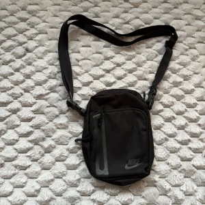 Svart axelväska från Nike - Säljer en svart axelväska från Nike med justerbar rem och flera fack. Väskan har en dragkedja framtill och en diskret logga i hörnet. Perfekt storlek för att bära med sig det viktigaste och passar till många olika outfits.