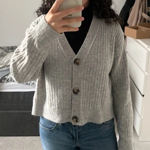 Grå ribbad kofta med stora knappar - Säljer en ljusgrå ribbstickad kofta från H&M med v-ringning och tre stora bruna knappar framtill. Koftan är croppad och har långa ärmar, perfekt att slänga på sig över en t-shirt eller topp. Superenkel att styla till jeans eller kjol.
