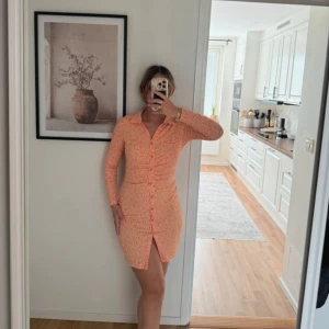 Kort orange klänning med knappar - Snygg kort klänning i orange med små vita prickar, långärmad och med knappar hela vägen framtill. Klänningen har krage och en liten slits nedtill. Perfekt för dig som gillar en figurnära passform och vill sticka ut.