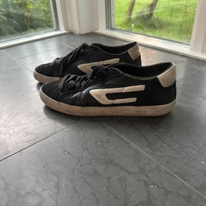 Svarta sneakers från Diesel - Säljer ett par svarta sneakers från Diesel med vita detaljer och logga på sidan. Skorna har snörning och platt sula. Dem är relativt slitna men fungerar fortfarande bra!
