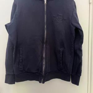 Mörkblå zip från GANT - Mörkblå hoodiejacka från GANT med dragkedja framtill och broderad logga på bröstet. Jackan har huva med snörning, ribbade muddar och två fickor på sidorna. Perfekt för en chill och stilren look.