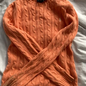 Kabelstickad orange tröja Ralph Lauren - Kabelstickad tröja från Ralph Lauren i en snygg orange färg. Klassisk rund halsringning och långärmad modell med broderad logga på bröstet. Tillverkad i 100% bomull, perfekt för lager-på-lager under höst och vår.