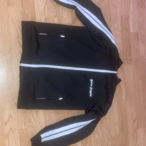 Palm Angels svart hoodie - Svart track jacket från Palm Angels med vita ränder längs ärmarna och vit dragkedja. Jackan har ståkrage, två fickor med dragkedja och logga broderad på bröstet. Perfekt för en sportig och trendig streetstyle-look.