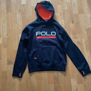 Svart hoodie Polo Sport Ralph Lauren - Svart hoodie från Polo Sport Ralph Lauren med stor vit och röd logga på bröstet. Insidan av huvan är orange mesh och tröjan har känguruficka framtill. Snygga orange detaljer vid ärmsluten. Perfekt för dig som gillar sportig och clean stil.