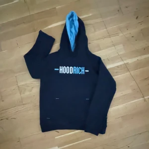 Svart Hoodie från Hoodrich med blå detaljer - Svart hoodie från Hoodrich med stor vit och blå logga på bröstet och mindre tryck på ryggen. Insidan av huvan är ljusblå, vilket ger en snygg kontrast. Klassisk känguruficka framtill och långa ärmar. Perfekt för dig som gillar streetwear vibes.