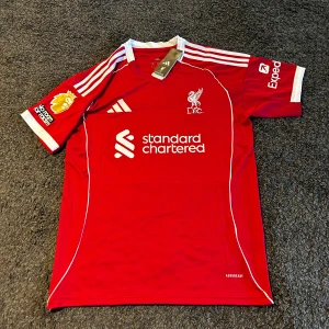 Liverpool röd matchtröja Adidas M - Liverpool FCs officiella röda matchtröja från Adidas med vita detaljer och broderat klubbmärke. Tröjan har korta ärmar, vita ränder på axlarna och tryck med sponsorloggor samt spelarnamn och nummer 7 på ryggen. Tillverkad i lätt och ventilerande polyester. Helt ny en replika