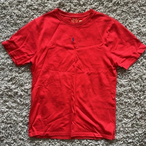 Röd t-shirt från Polo Ralph Lauren - Snygg röd t-shirt från Polo Ralph Lauren i storlek XL. Klassisk rund hals och korta ärmar. På bröstet finns den ikoniska blå broderade Polo-loggan. Tillverkad i mjuk bomull som är skön mot huden. Perfekt för dig som gillar stilrena och tidlösa plagg.