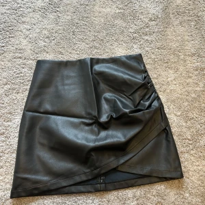 Svart omlottkjol i PU - Snygg svart kortkjol i PU-material med omlott-design och lätt rynkning på sidan. Kjolen har en stilren look och passar perfekt till en cool outfit. Modellen är rak och har en diskret dragkedja bak för enkel på- och avtagning. Står strl L men passar på M