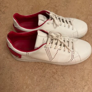 Valentino Garavani vita sneakers - Säljer ett par snygga vita Valentino Garavani sneakers med röda detaljer och nitar på hälen. Skorna har perforerat V-mönster på sidan, rund tå och platt sula. Insidan är röd och de har klassisk snörning. Perfekta för dig som gillar lyxiga och stilrena sneakers.