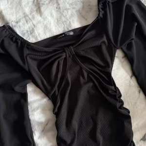 Svart långärmad klänning från SHEIN - Supersnygg svart kortklänning från SHEIN med puffiga långärmade ärmar och ribbad struktur. Klänningen har en cool twistad detalj framtill och smickrande rynkningar längs sidorna. 