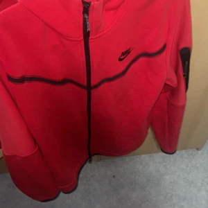Röd Nike hoodie med dragkedja M - Nike hoodie i rött med hel dragkedja och svart logga på bröstet. Snygga svarta detaljer och en praktisk ficka med dragkedja på ärmen. Huva och långa ärmar gör den perfekt för chill eller träning.