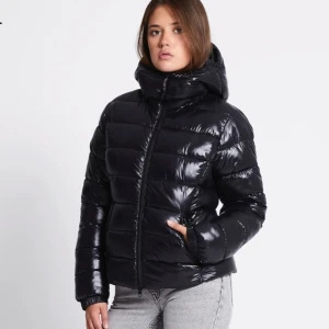 Svart glansig pufferjacka med huva - Säljer en svart, glansig pufferjacka med huva och dragkedja. Jackan är vadderad och har en schysst oversized look, perfekt för kalla dagar. Den har långa ärmar och breda kanaler som ger en cool och modern vibe.