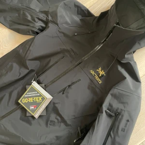 Svart Arc'teryx Gore-Tex vindjacka M - Säljer en svart vindjacka från Arc'teryx i storlek M. Jackan är tillverkad i slitstark Gore-Tex och har huva, flera dragkedjeförsedda fickor och snygg gul logga på bröstet. Perfekt för dig som vill ha en stilren och funktionell jacka med grym passform.