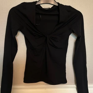 Svart långärmad topp  - Svart långärmad topp från Monki i storlek XS. Toppen har en snygg twistad detalj framtill vid bysten och djupare urringning. Tillverkad i mjuk bomullsmix, perfekt för att lyfta din outfit med en cool touch.