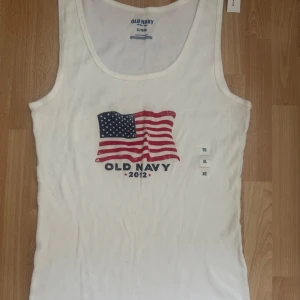  Ny. Vit ribbad linne från Old Navy XL - Vit ribbad linne från Old Navy med amerikansk flagga och tryck 'OLD NAVY 2012' framtill. Ärmlös modell med rund halsringning, perfekt för sommardagar. Mjuk bomullskänsla och klassisk passform. Köpte i USA. Tryckt är avsiktligt sprucken.