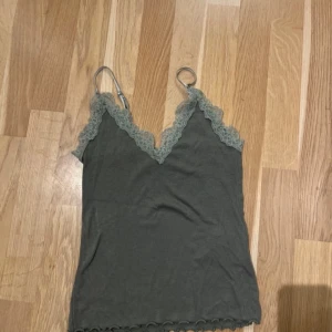 Grå linnetopp med spetskant - Supersnygg grå linnetopp med tunna axelband och v-ringad spetskant i ljusgrönt, hela toppen är ljusare än på bild. Toppen har en croppad passform och vågig kant nedtill. Perfekt att styla med jeans eller kjol för en chill look. 