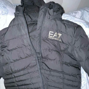 Svart EA7 dunjacka med huva - Säljer en svart dunjacka från EA7 Emporio Armani med huva och dragkedja framtill. Jackan har ett stilrent, quiltat mönster och logga på bröstet. Perfekt för dig som vill ha en snygg och varm jacka med exklusiv känsla.