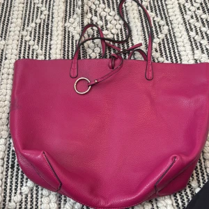 Rosa shoppingväska i skinn - Stilren och rymlig shoppingväska i stark rosa färg med svarta detaljer. Väskan är tillverkad i mjukt skinn och har dubbla handtag samt en dekorativ metallring framtill. Insidan är svart och öppen utan fack, perfekt för att slänga ner allt du behöver.