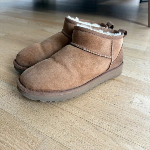 Bruna UGG boots i mocka med fårskinn - Mysiga bruna boots från UGG i mjuk mocka med fluffigt vitt fårskinnsfoder. Skorna har rund tå, platt sula och dragflik bak. Perfekta för kalla dagar och ger en chill vibe till din outfit. De är 100% äkta.
