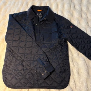 Mörkblå quiltad jacka från Morris - Quiltad mörkblå jacka från Morris med klassisk krage i manchester, bröstficka med knapp och snyggt rutmönster. Jackan har tryckknappar framtill och en patch med brittisk flagga på ärmen. Perfekt för dig som gillar stilren och tidlös look.