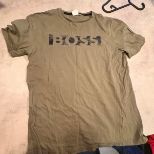 Olivgrön BOSS t-shirt med tryck - Snygg olivgrön t-shirt från BOSS i storlek M. T-shirten har ett svart BOSS-tryck på bröstet och klassisk rund hals. Modellen är kortärmad och gjord i mjuk bomull, perfekt för en chill och stilren look.