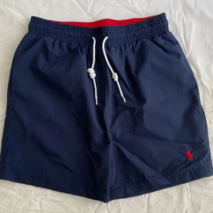 Polo Ralph Lauren Shorts  - Snygga marinblå badshorts från Polo Ralph Lauren med röd insida i midjan och vit snörning framtill. Klassisk Polo-logga broderad i rött på benet. Shortsen har elastisk midja och en bakficka. Använda enbart en gång. Storlek M men passar mer för S. Priset kan diskuteras 😊