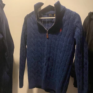 Blå stickad tröja från Polo Ralph Lauren - Säljer en snygg blå stickad tröja från Polo Ralph Lauren med dragkedja och det klassiska röda logotypbroderiet på bröstet. Tröjan har en ribbad krage och långa ärmar, perfekt för kyliga dagar. Passar både till vardags och mer uppklädda tillfällen.