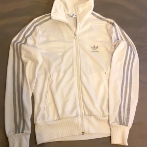 Vit Adidas track jacket med silverränder - Snygg vit Adidas track jacket med klassiska silverfärgade ränder längs ärmarna och stor Trefoil-logga på ryggen. Hög krage, hel dragkedja och två fickor framtill. Perfekt för en sportig och clean look. Tröjan är liten i storleken trots att det står 42 på tröjan jag tycker att det är stolek S,på armen finns litet håll som syns på bilden.