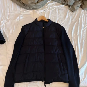 Svart pufferjacka med dragkedja - Snygg svart pufferjacka med quiltade paneler framtill och släta ärmar. Jackan har en stilren look med ståkrage och dragkedja hela vägen. Perfekt för dig som vill ha en enkel men modern jacka till höst och vinter.