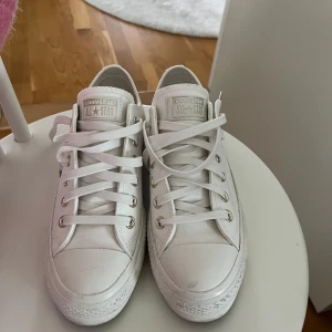 Vita Converse All Star sneakers - Säljer ett par klassiska vita Converse All Star sneakers med lågt skaft och platt sula. Skorna har rund tå, snörning med metallhål och tydlig logga bak på hälen. Materialet är slitstark canvas och gummisula, perfekt för dig som gillar clean och enkel stil. Storlek 37,5 