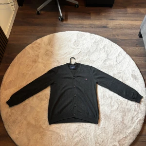 Mörkgrå kofta från Bondelid - Säljer en stilren mörkgrå kofta från Bondelid med v-ringning och knappar framtill. Klassisk design med lång ärm och liten logga på bröstet. Perfekt att slänga på sig över en t-shirt eller skjorta för en clean look.