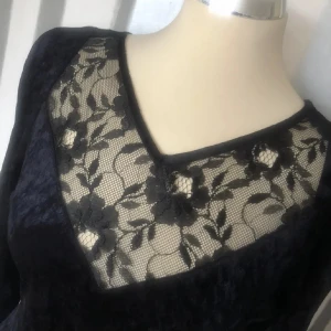 Svart sammetsklänning med spetsdetaljer - Elegant svart sammetsklänning med lång ärm och v-ringning. Bröstpartiet och ärmsluten har transparent spets med blommigt mönster som ger en lyxig känsla. Klänningen är figurnära och har en klassisk siluett som passar perfekt när du vill känna dig extra snygg.