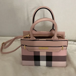 Rutig rosa handväska med axelrem - Säljer en snygg handväska i  ljusrosa med klassiskt rutigt mönster i svart, vitt och rosa. Väskan har både handtag och avtagbar axelrem i matchande färg samt guldfärgade detaljer. Säljer på grund av att den inte kommer till användning. Kontakta för flera frågor 🩷 den är köpt för 500 och säljer den för samma pris. 