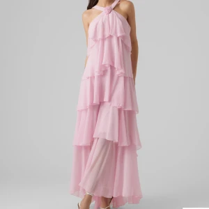 Ljusrosa långklänning med volanger - Ny pris är 1199kr. Från Vero Moda. Säljer en drömmig ljusrosa långklänning med flera lager volanger och omlottband i halsen som bildar en rosdetalj. Klänningen har öppen rygg och breda axelband, vilket ger en elegant och romantisk vibe. Materialet är tunt och flowy, perfekt för sommarens speciella tillfällen.