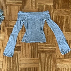 Grå offshoulder topp från Gina Tricot - Superfin ljusgrå offshoulder topp från Gina Tricot i mjukt och stretchigt material. Toppen har långa ärmar och en croppad passform med vågig kant nertill. Perfekt att styla med jeans eller kjol för en trendig look.