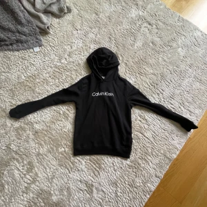 Svart hoodie från Calvin Klein Jeans - Svart hoodie från Calvin Klein Jeans med vit logga på bröstet. Klassisk modell med huva och långa ärmar. Tillverkad i 100% bomull för en skön och mjuk känsla. Perfekt för en chill och stilren look.