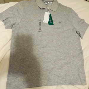 Grå pikétröja från Lacoste XL - Snygg grå pikétröja från Lacoste i regular fit. Klassisk krage, knappar framtill och diskret Lacoste-logga på bröstet. Tillverkad av bomull och polyester med lite elastan för extra komfort. Perfekt för en clean och stilren look.
