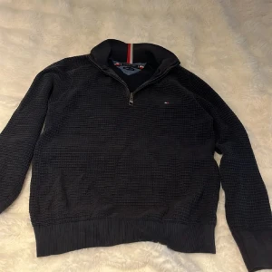 Svart half zip tröja Tommy Hilfiger - Svart långärmad tröja från Tommy Hilfiger i ekologisk bomull. Tröjan har ett snyggt rutmönster, ribbade muddar och krage med half zip och röd-vit detalj. Liten logga på bröstet. Perfekt till chill eller skola.