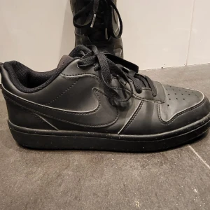 Svarta Nike sneakers i skinn - Snygga helsvarta Nike sneakers i skinn med klassisk swoosh på sidan. Lågt skaft, snörning och vadderad plös för extra komfort. Perfekta för dig som gillar en clean och stilren look till vardags. Platt sula och rund tå. Har använts få gånger under hösten. Kan mötas upp I Stockholm.