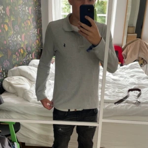 Grå pikétröja från Polo Ralph Lauren - Säljer en stilren grå långärmad pikétröja från Polo Ralph Lauren. Tröjan har klassisk krage, knappar vid halsen och den ikoniska broderade loggan på bröstet. Perfekt för dig som gillar en clean och tidlös look. Skick 8/10