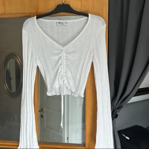 Vit croppad ribbad topp med snörning - Superfin vit croppad topp från Fashion med långa ärmar och v-ringning. Toppen har ribbat mönster, snygga håldetaljer och snörning framtill som ger en cool rynkad effekt. Perfekt för dig som gillar trendiga och feminina plagg.