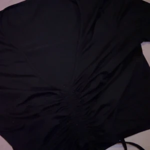 Svart tröja med snörning - Snygg svart tröja med djup v-ringning och smickrande snörningsdetalj i midjan. Tröjan är perfekt för dig som gillar en stilren look med en twist.